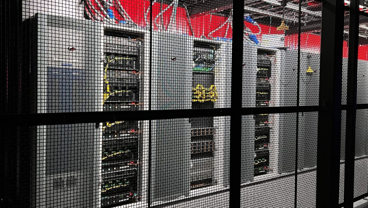 Server racks with network cables and switches inside a data center, viewed through black mesh security enclosure.