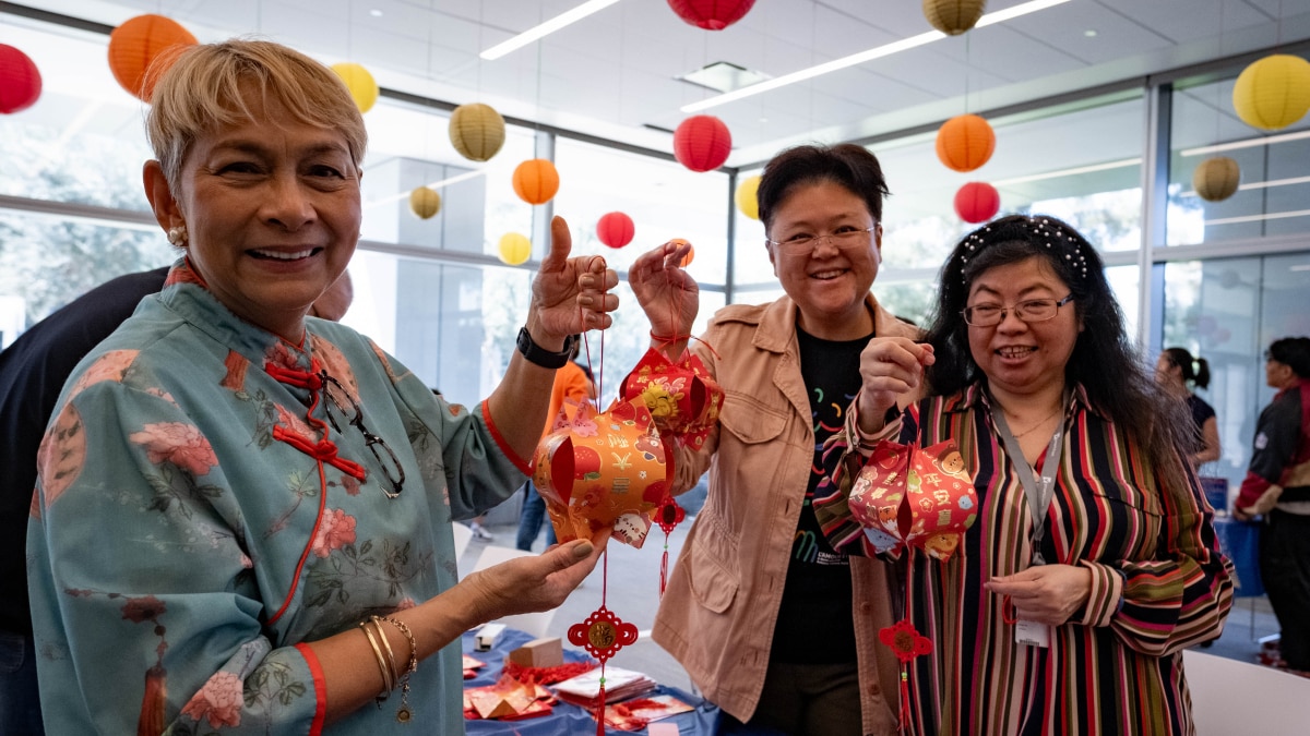 Three women smiling indoors, holding handmade red and orange paper lanterns with hanging decorations; festive lanterns overhead.