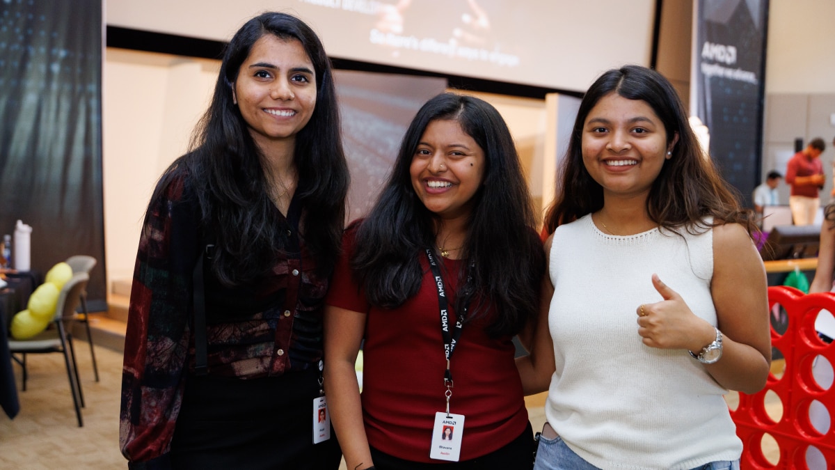 Three women smiling at a corporate event, wearing badges with AMD lanyards in a modern workspace.