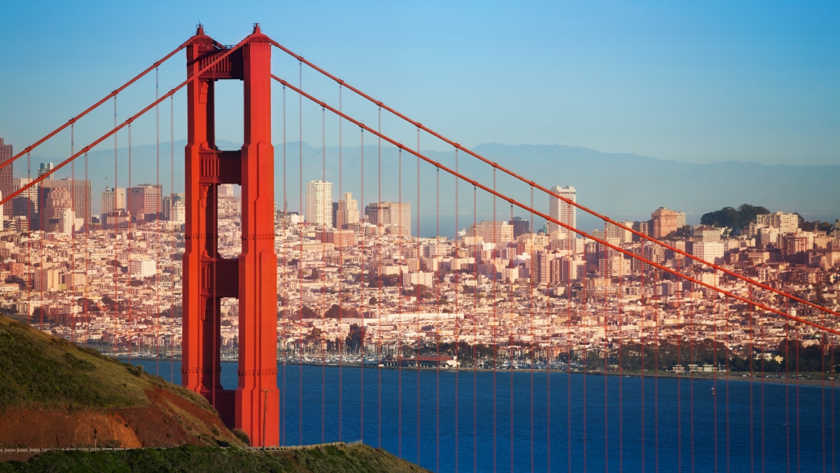 Golden Gate Bridge tower with red cables spanning blue bay and city skyline backdrop.