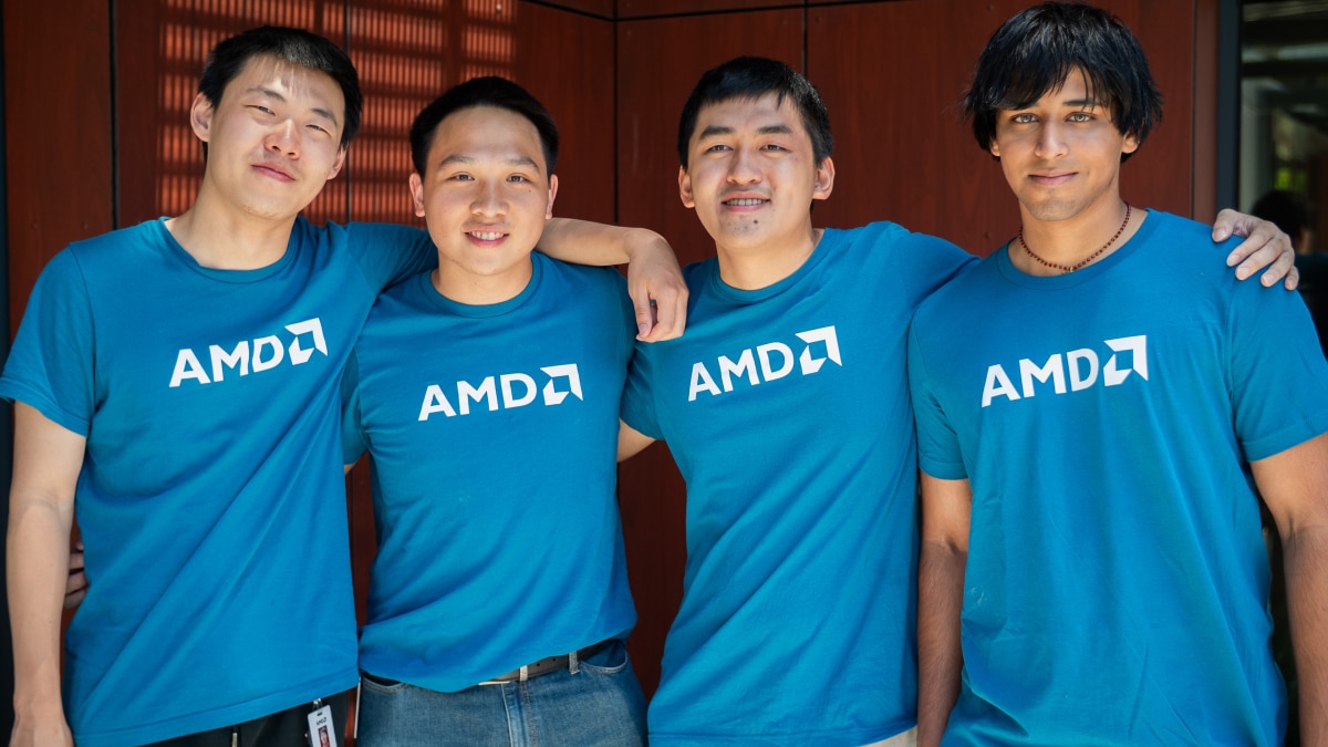 Four people wearing blue AMD shirts standing together with arms around each other, smiling.