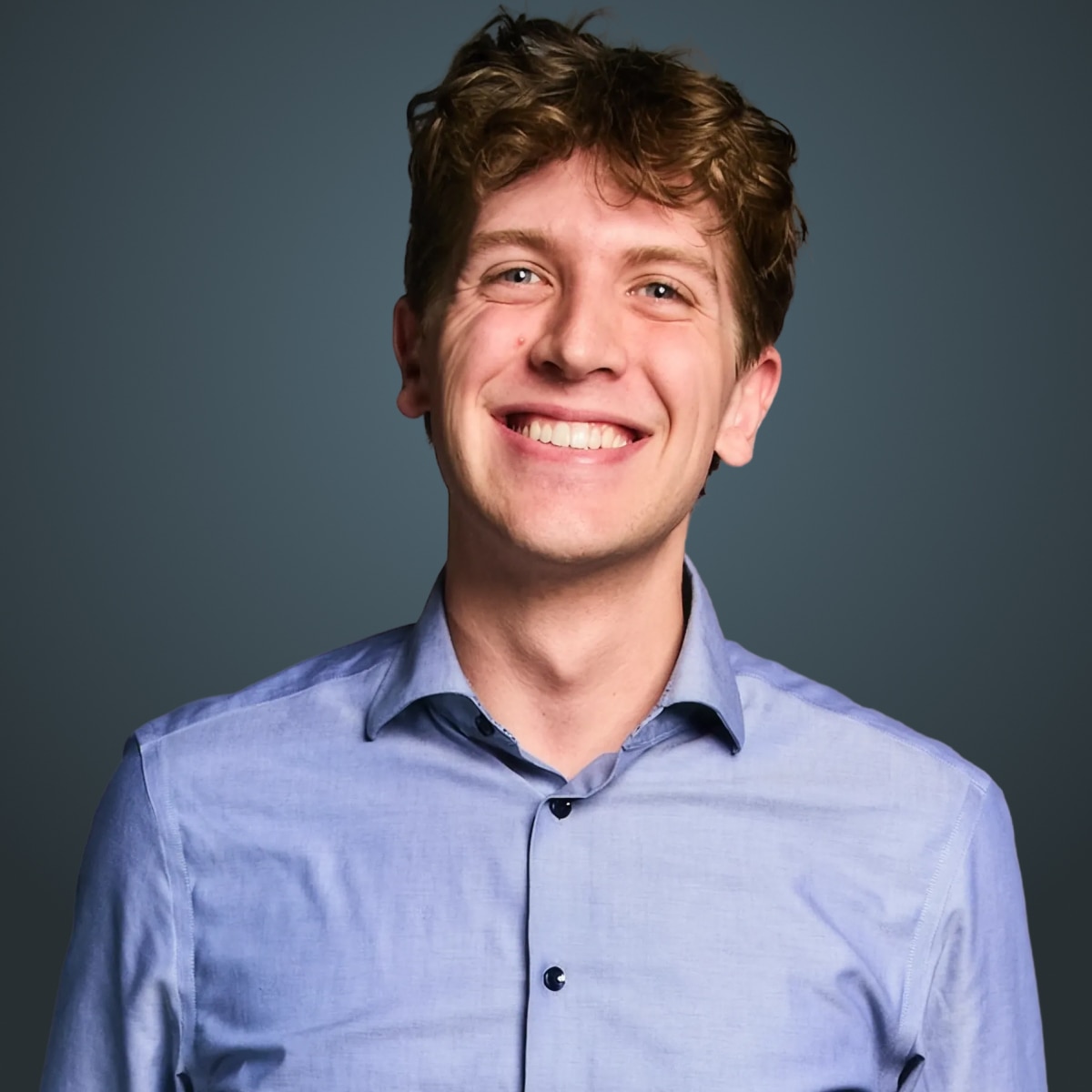 Smiling person with short wavy brown hair wearing a light blue button-down shirt against a dark teal background.