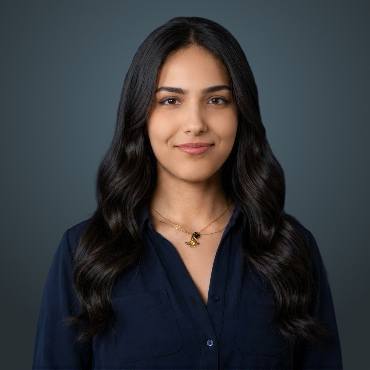 Person with long dark wavy hair wearing a navy blouse and gold necklace against a dark teal background.