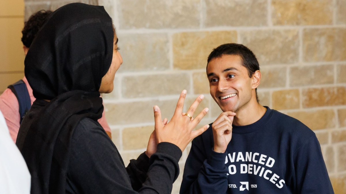 Two students smile and converse indoors, one gesturing while the other wears an Advanced Micro Devices sweatshirt.