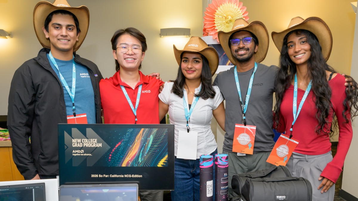 Five New College Grad Program attendees pose together at an event booth with hats, badges, laptops, and giveaways.