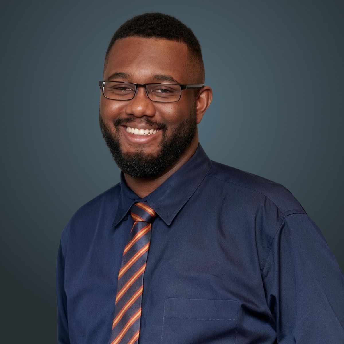 Smiling person with glasses and a beard wearing a navy shirt and striped tie against a dark teal background.