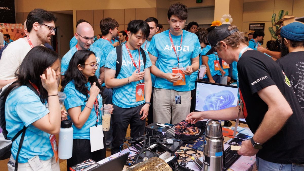 Attendees in blue shirts gather around a technology demo table with computer hardware and a monitor at an expo.