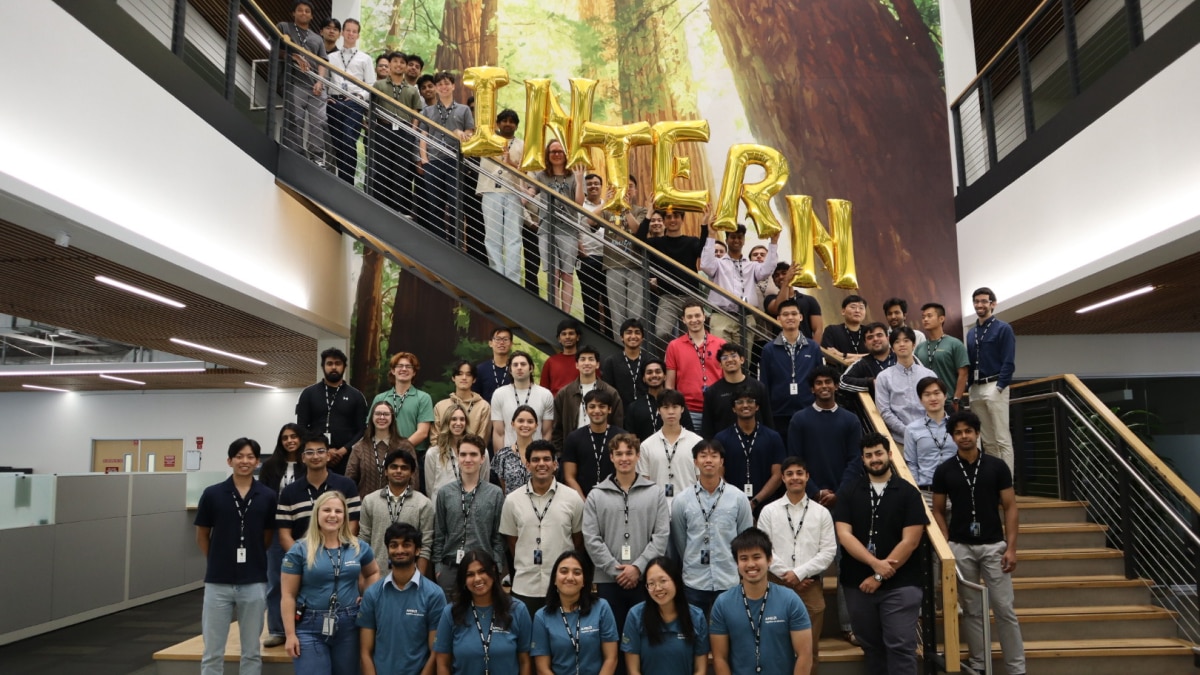 Large group of interns poses on and around an office staircase beneath gold balloon letters spelling “INTERN.”
