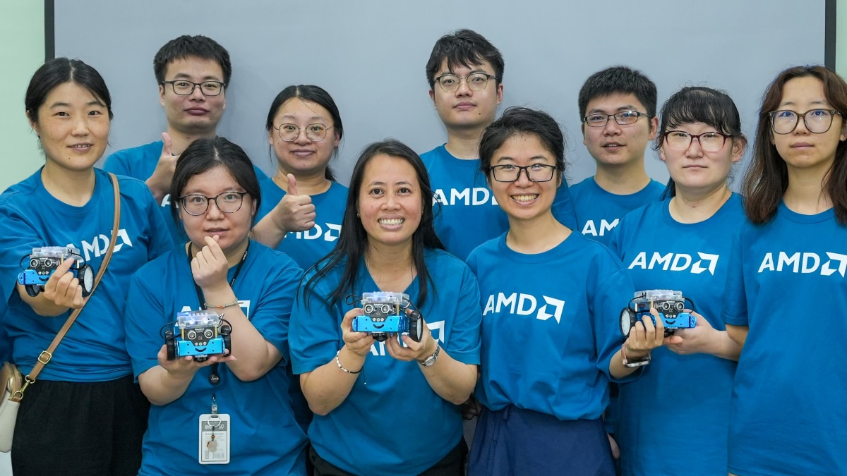 Group of people in blue AMD shirts pose indoors, several holding small educational robots and smiling at the camera.