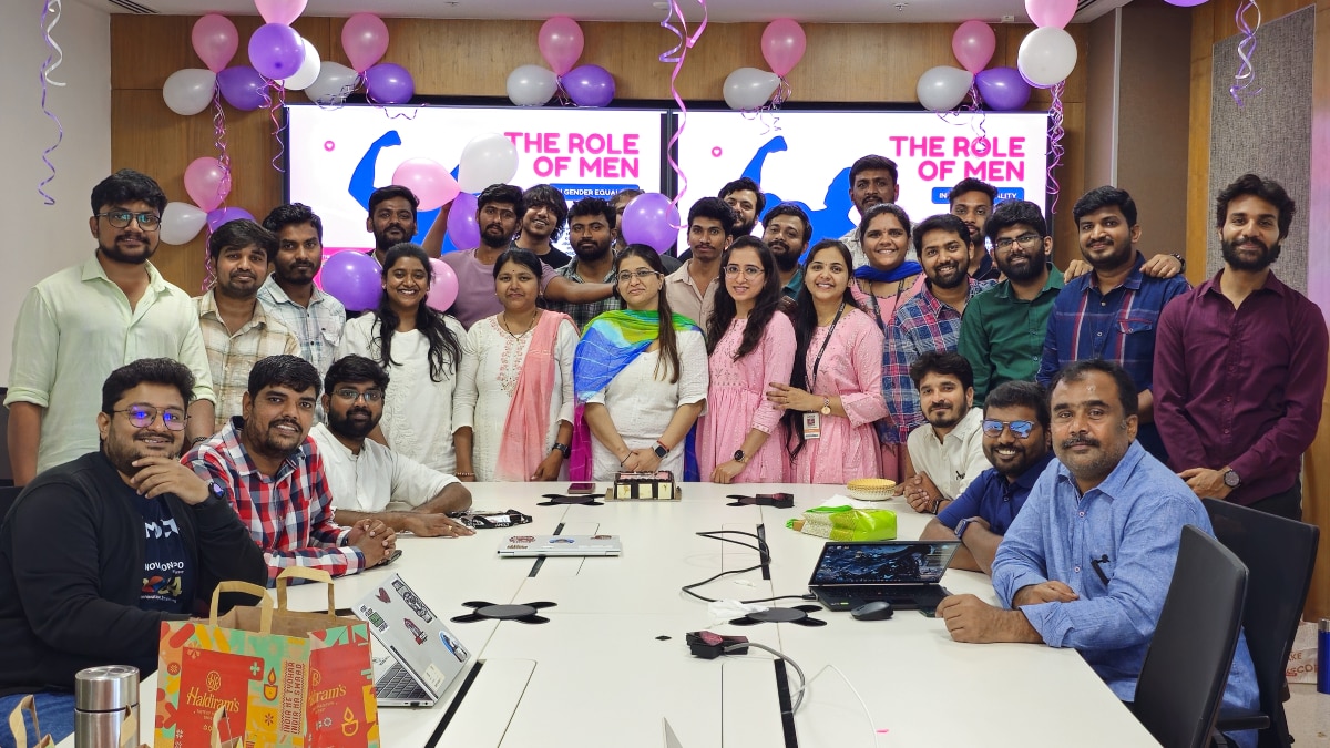 Team posing in a conference room decorated with pink, purple, and white balloons; screen reads "THE ROLE OF MEN."