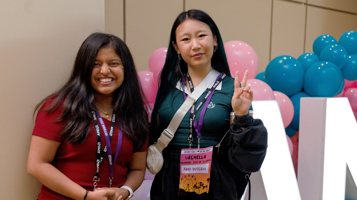 Two AMD interns wearing event badges smile and pose in front of pink and blue balloons, with one making a peace sign.