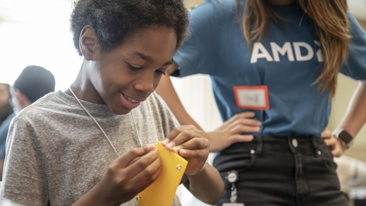Student smiles while assembling a small project during a hands-on STEM activity, with AMD volunteers in the background.