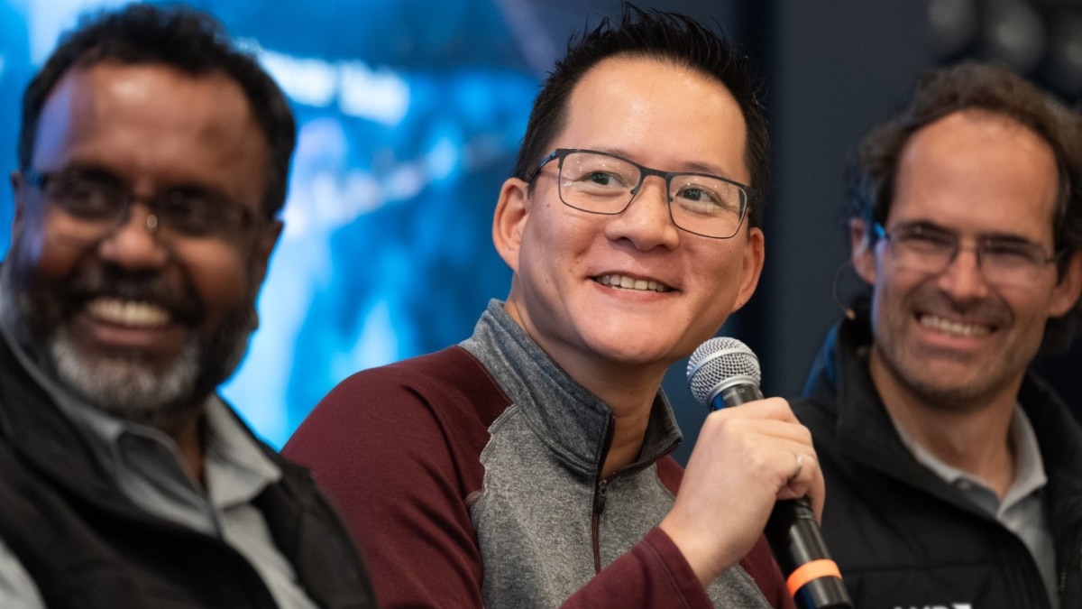 Three speakers smile during a panel discussion, with the center speaker holding a microphone while addressing the audience.