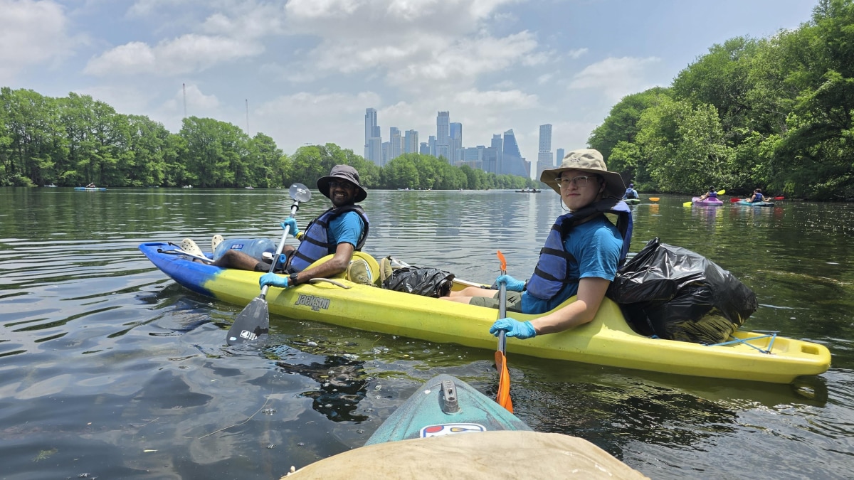 Two volunteers in a tandem kayak collect trash on a river while wearing life jackets and gloves, with a city skyline in the background.