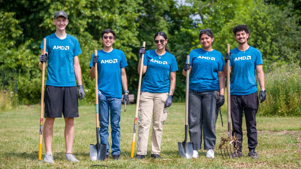 Five volunteers in blue AMD T-shirts stand outdoors holding shovels and a rake, ready for a community cleanup event.