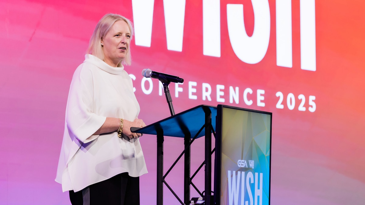 Speaker stands at a podium delivering a presentation at a 2025 conference, with a microphone and large conference backdrop in the background.