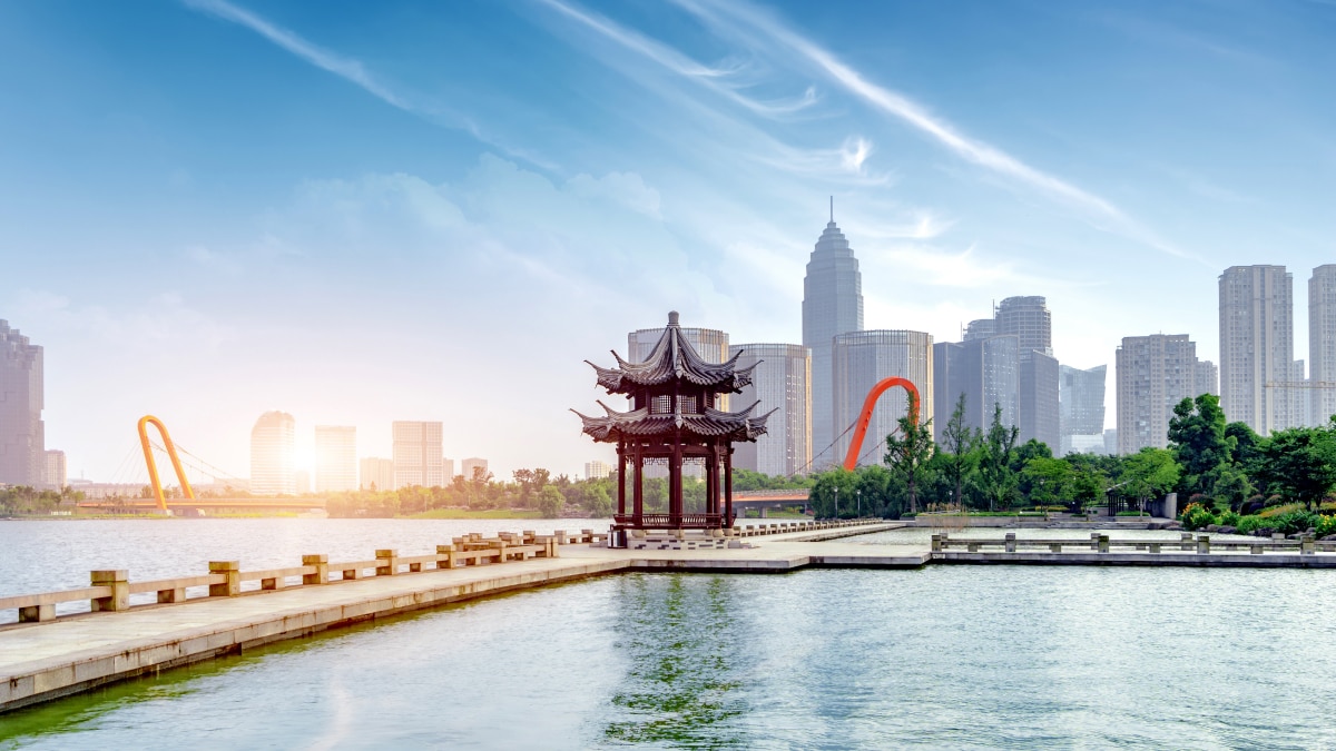 A scenic view of a traditional Chinese pavilion on a lake walkway, with a modern city skyline and a distinctive orange bridge in the background under a bright sky.