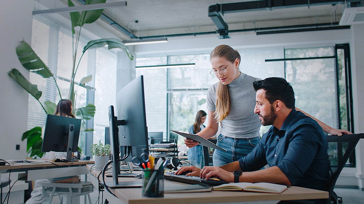 Two coworkers collaborating in a modern office; a woman holding a tablet is discussing something with a man working on a desktop computer.