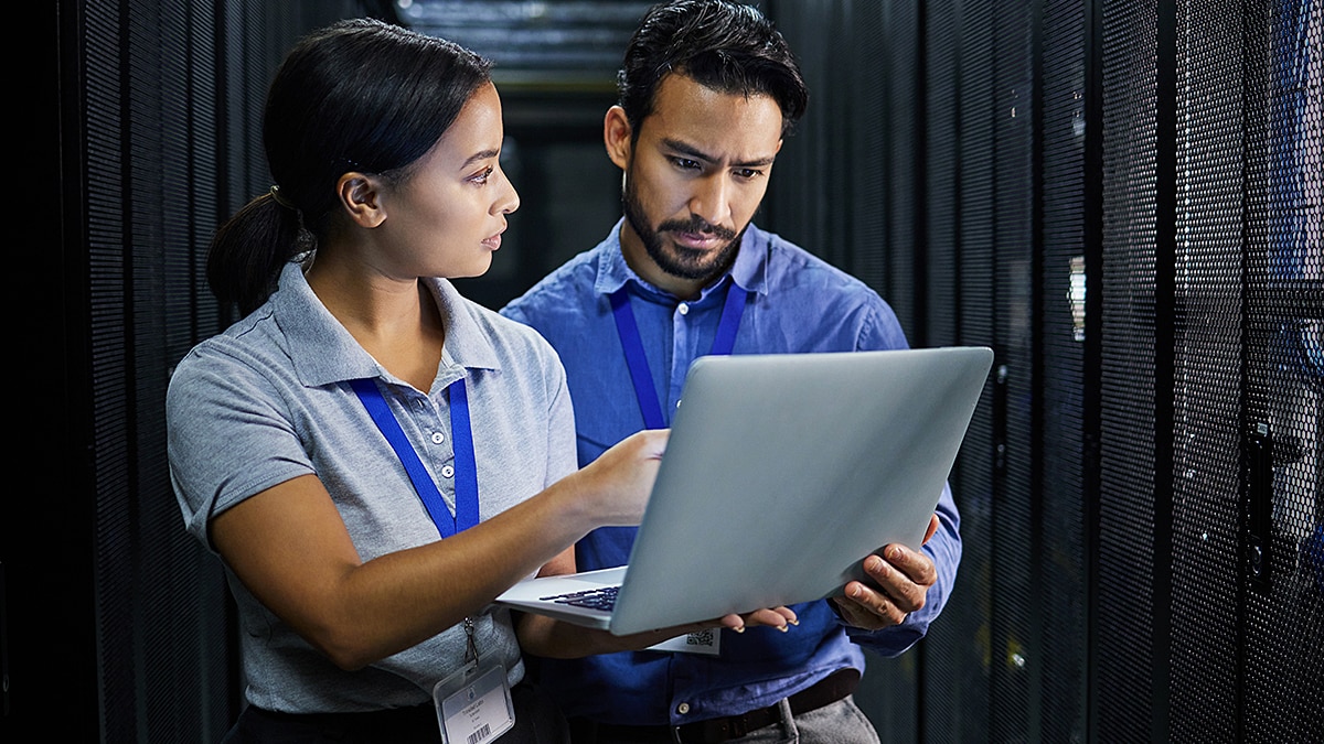 Two IT professionals standing in a data center aisle, discussing while looking at a laptop computer.