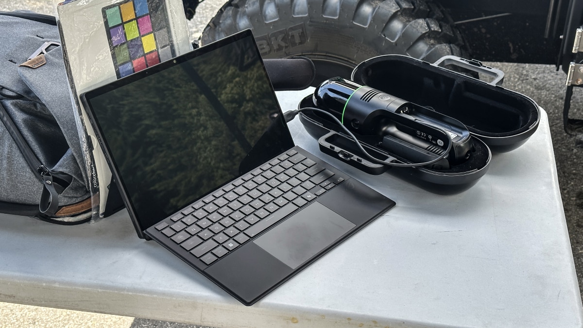 Open laptop on a table beside a handheld 3D scanner in a black case, with a backpack and color calibration chart nearby.