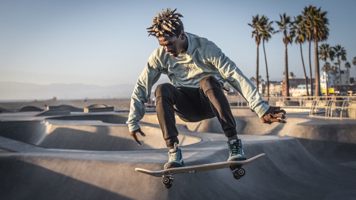 Skateboarder in mid-air performing a trick at a concrete skatepark with palm trees in the background.