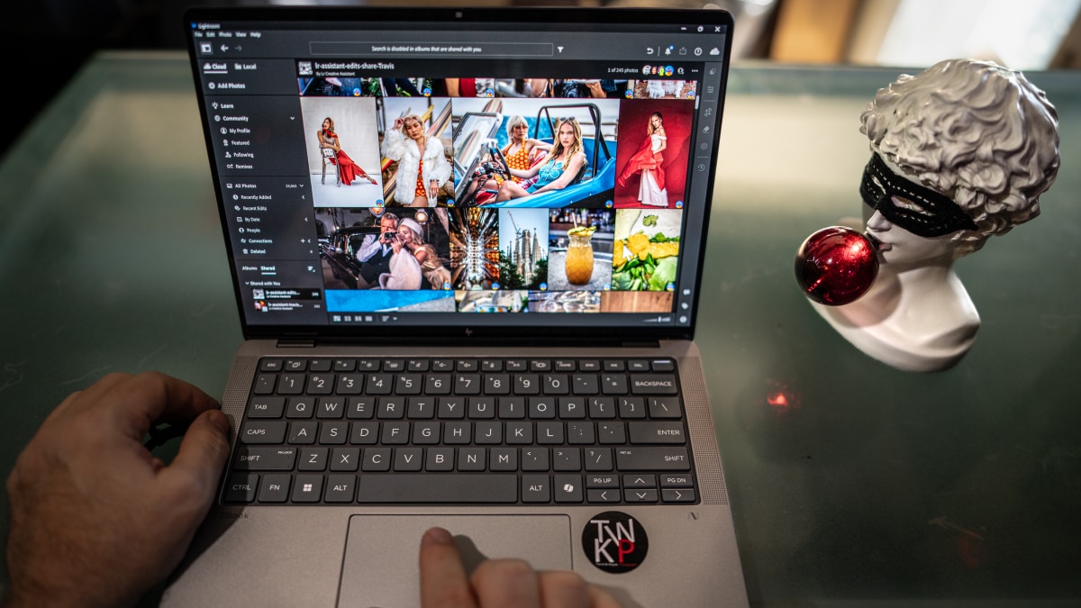Laptop screen displaying photo editing software with fashion images, hand on keyboard, bust sculpture beside it.