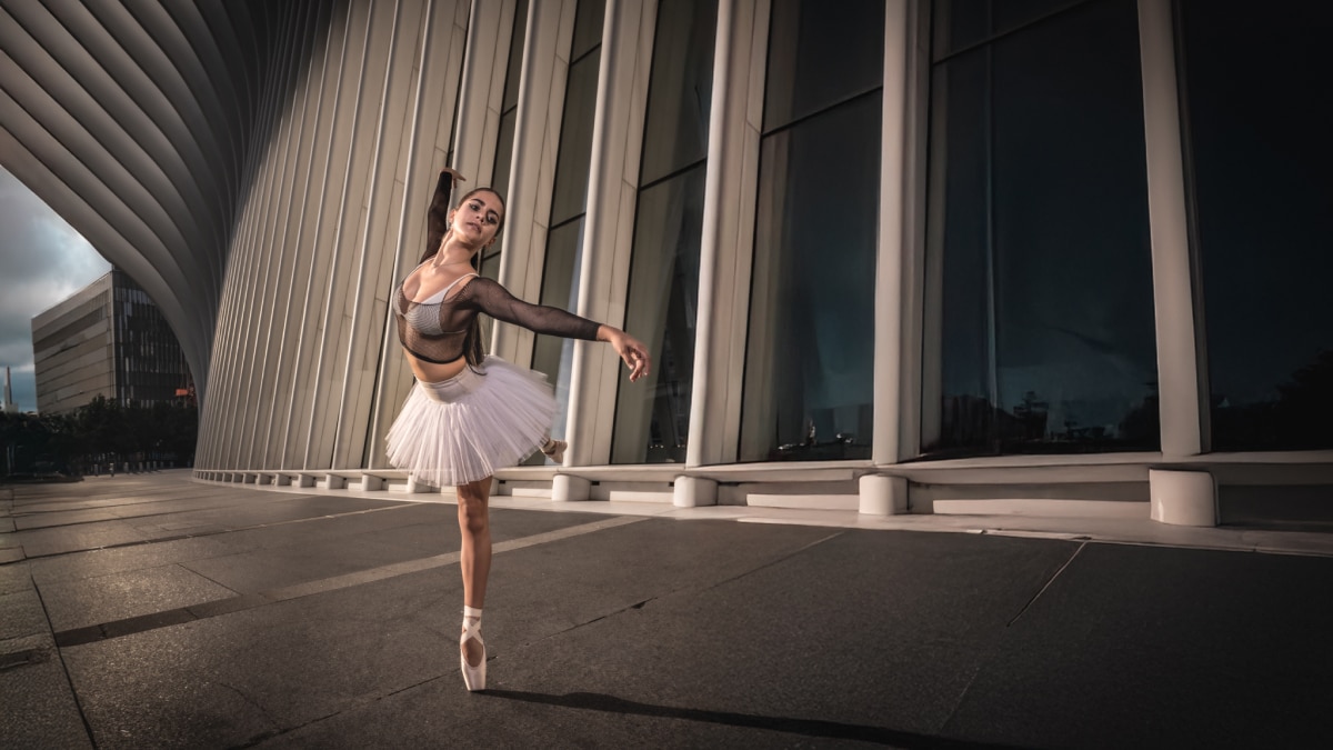 Ballet dancer in white tutu and black mesh top balancing en pointe outside modern architectural building.