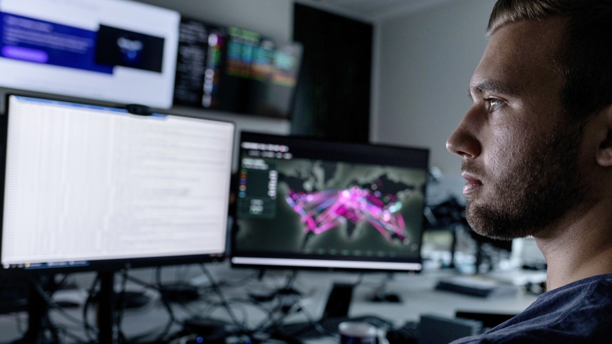 Analyst looks at two monitors showing code and a world map with bright network visualization lines.