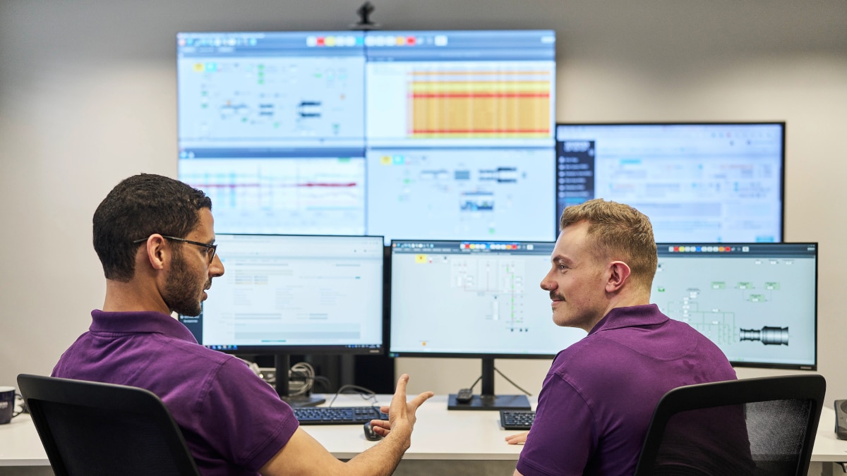 Two engineers in purple shirts discuss data while monitoring multiple computer and wall screens in control room.