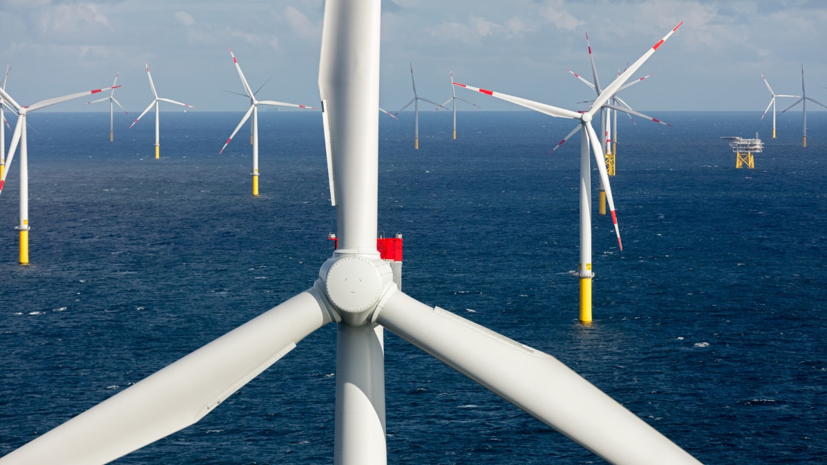 Large offshore wind turbines with yellow bases rotating over deep blue sea under partly cloudy sky.