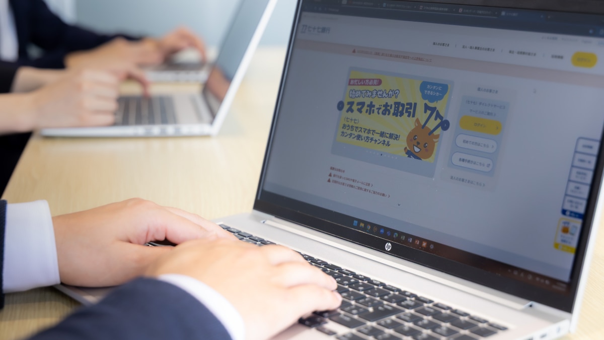 Person typing on a laptop displaying a 77 Bank webpage with illustrated character and service buttons.