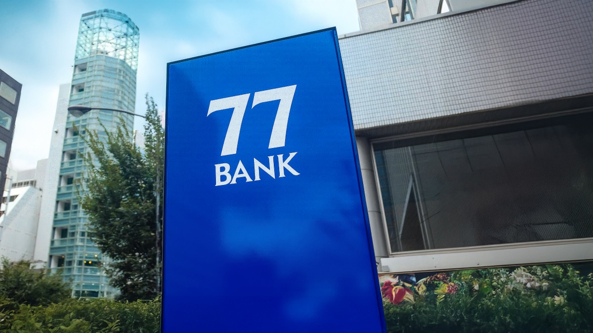 Blue outdoor sign displaying the white “77 BANK” logo with city buildings in the background.