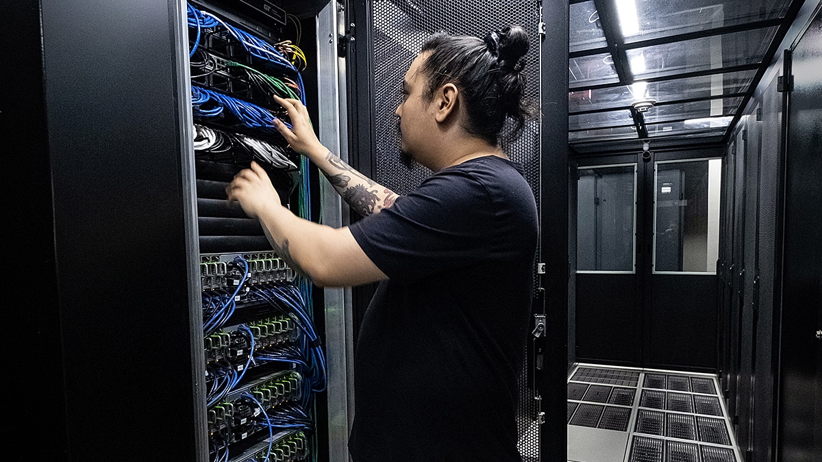 Technician adjusting blue and black network cables in a server rack inside a data center aisle.