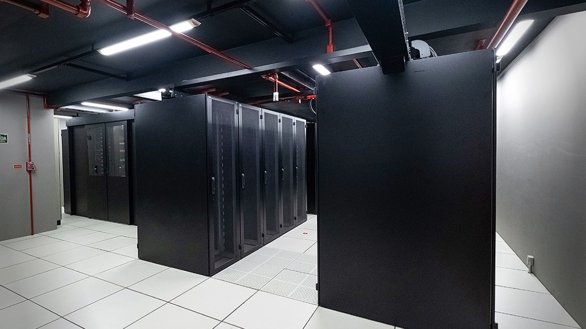 Row of black server racks in a clean, brightly lit data center with white tiled floor and ceiling pipes.