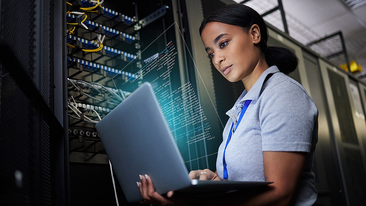 Technician in server room analyzing code on laptop with glowing data overlay.