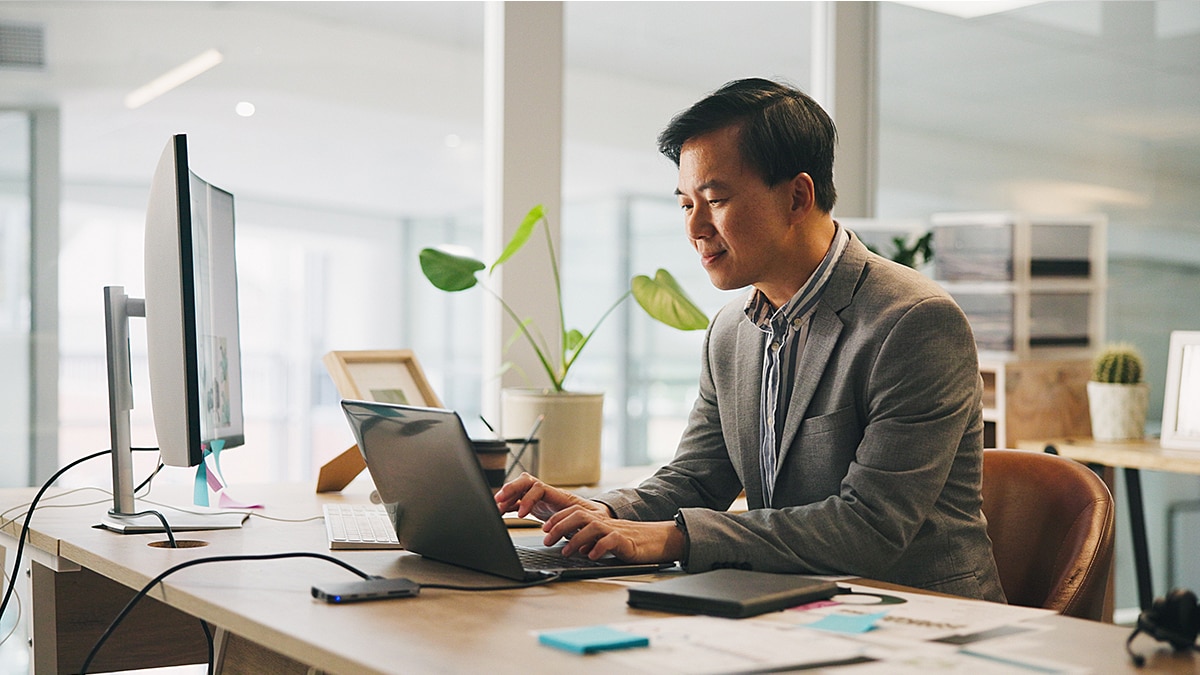 Business professional typing on laptop at desk with monitor and indoor plants.