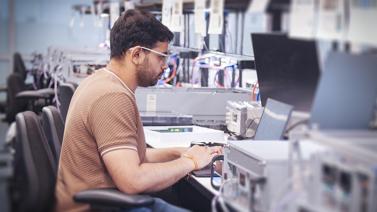 Engineer seated at a workstation using a laptop beside test equipment and network hardware in a lab.