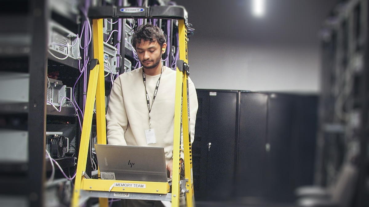 Technician using a laptop on a yellow ladder shelf labeled “Memory Team” surrounded by server racks and cables.