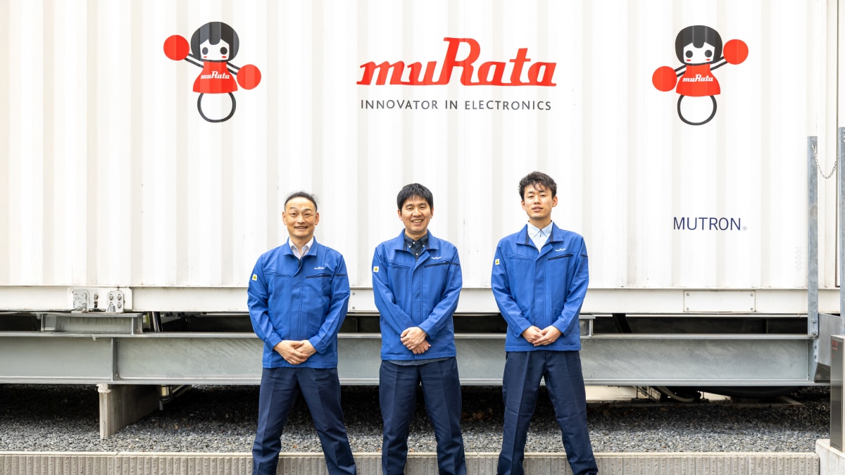 Three engineers in blue uniforms standing before muRata logo wall with cartoon mascot graphics.