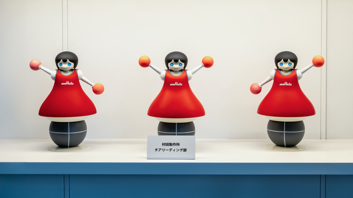 Three red muRata cheerleader robots with black hair and pom-poms displayed on a white shelf.