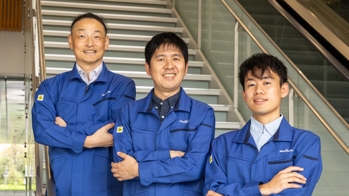 Three muRata engineers in blue uniforms posing confidently on staircase indoors.