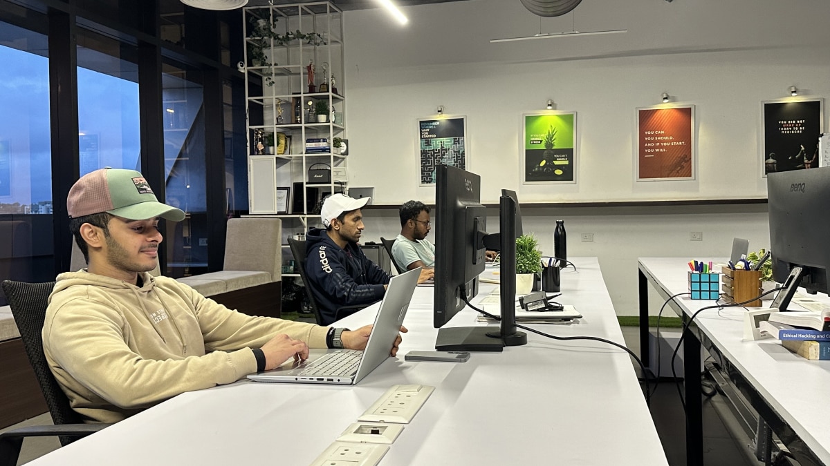 Three people working at a long office desk with laptops and monitors, surrounded by posters and shelving.
