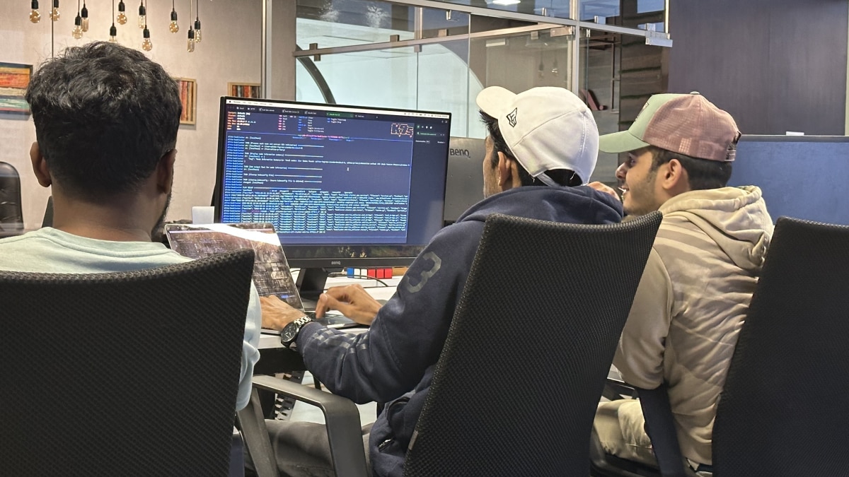 Three developers seated at a desk reviewing code displayed on a large monitor, with laptops and office decor around them.