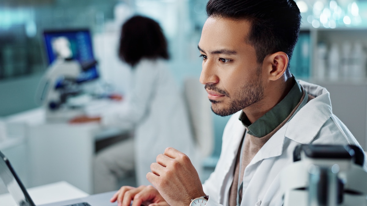 Researcher focuses on a computer in a lab with a microscope and blurred colleague in background.