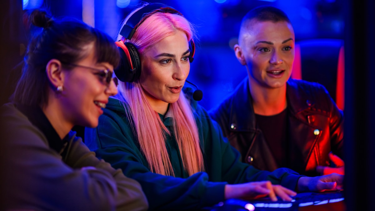 Three women focused on a game at a PC, wearing headsets in a blue and pink neon-lit gaming area.