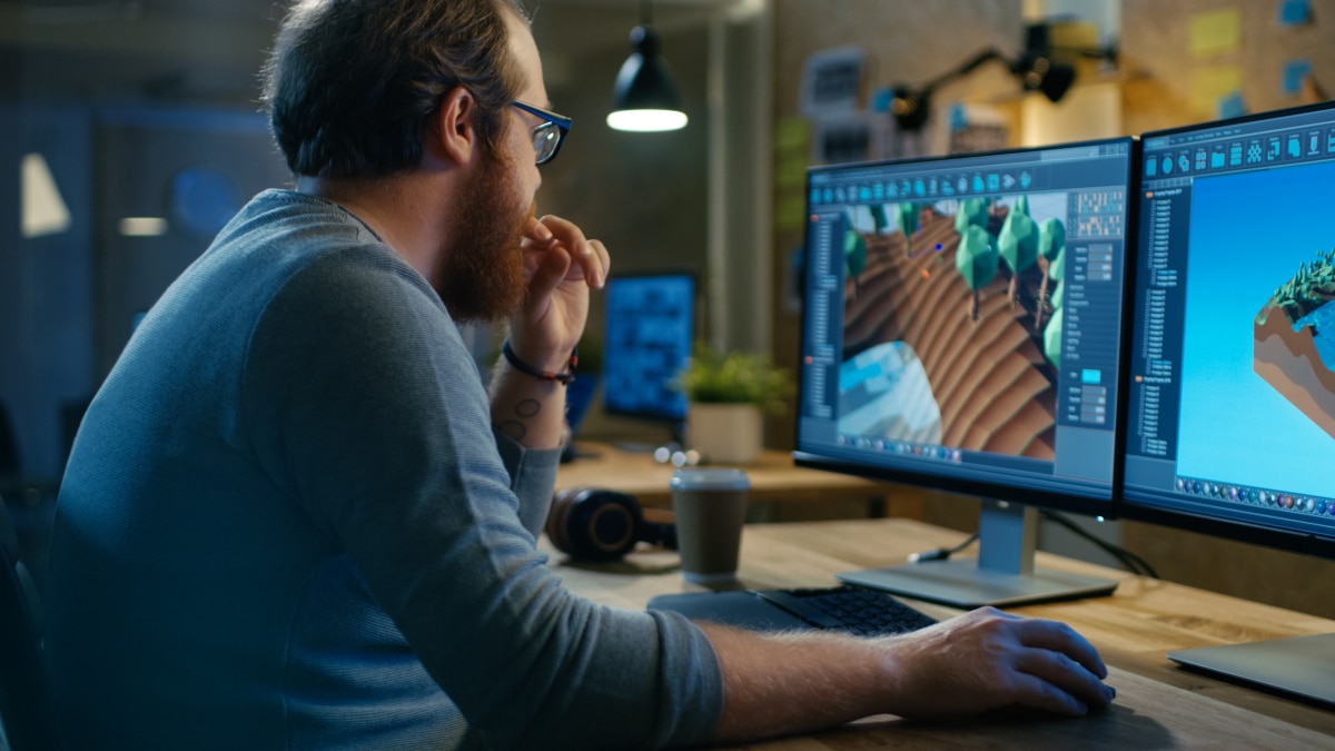 Developer at dual monitors editing a 3D terrain scene in modeling software in a dim workspace.