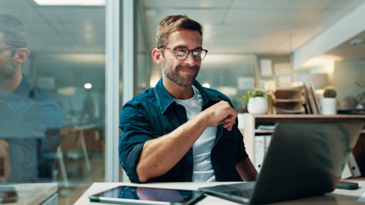 Professional wearing glasses works on a laptop at a desk in a glass-walled office with tablet and documents nearby.