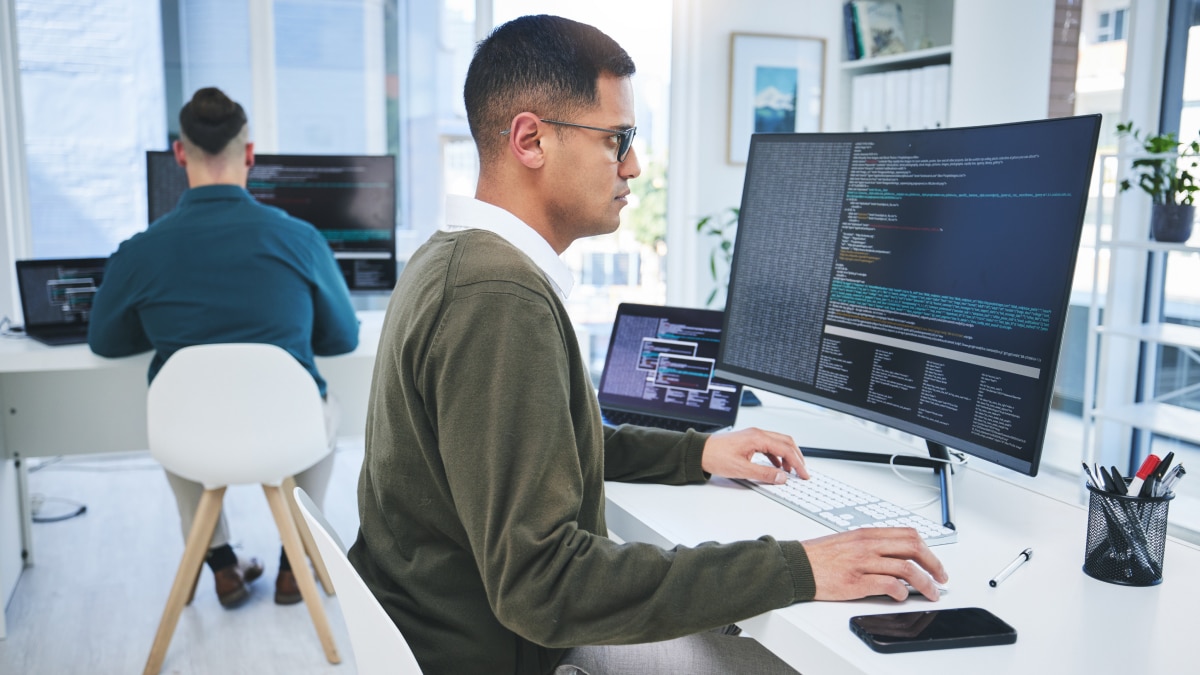 Two developers working at desks with large monitors showing code in a bright, modern office environment.