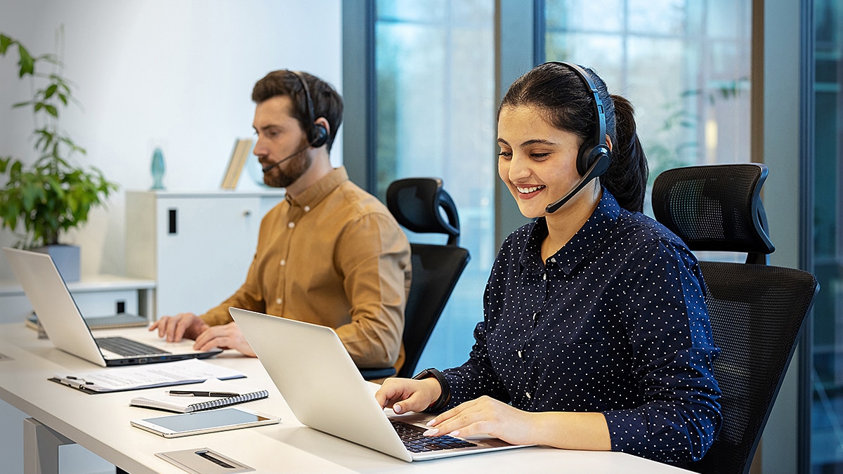 Two customer support agents wearing headsets work on laptops in a modern office with blue lighting.