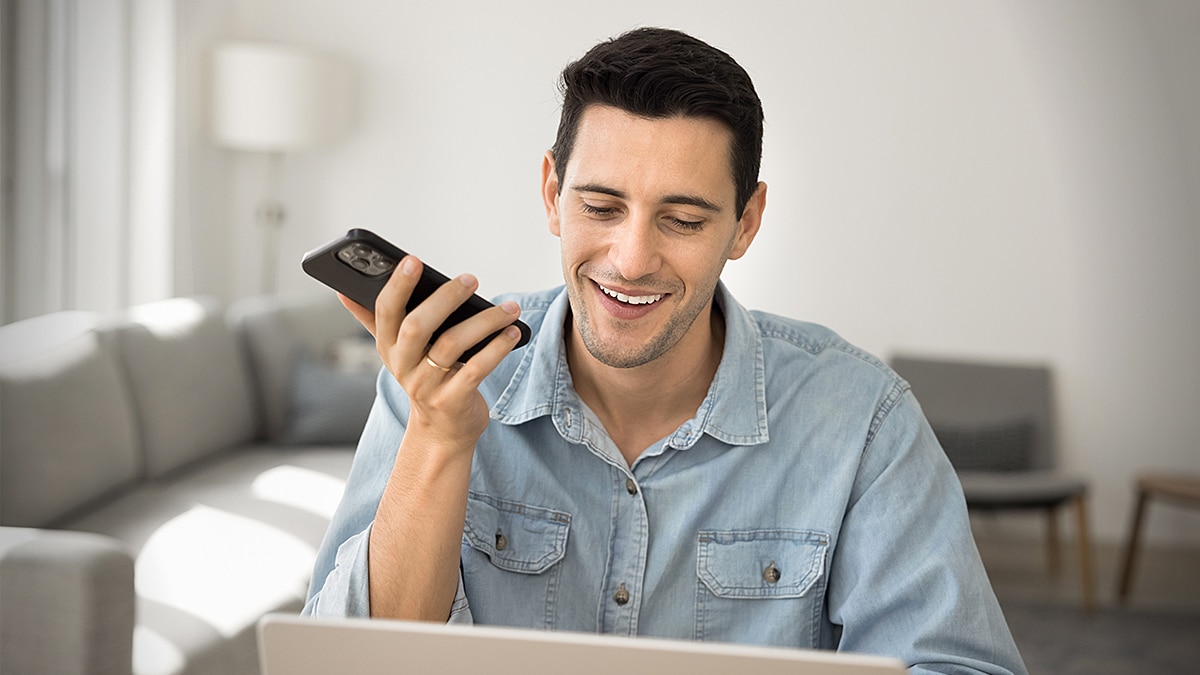 Man smiling while speaking into a smartphone, seated with a laptop in a bright home living room.